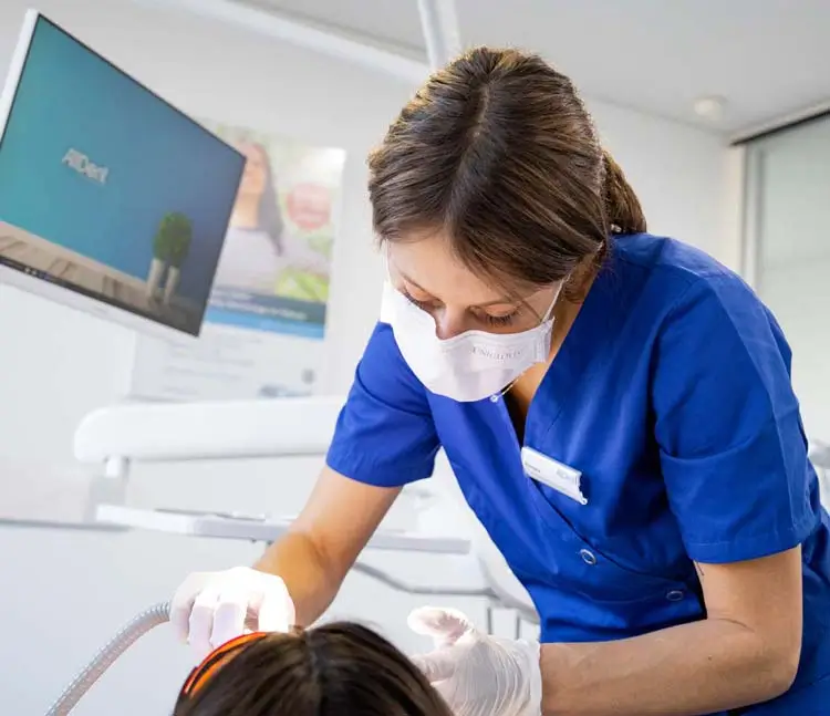 Zahn&auml;rztin in blauer Arbeitskleidung und Maske behandelt eine Patientin im Behandlungsstuhl, im Hintergrund moderner Bildschirm