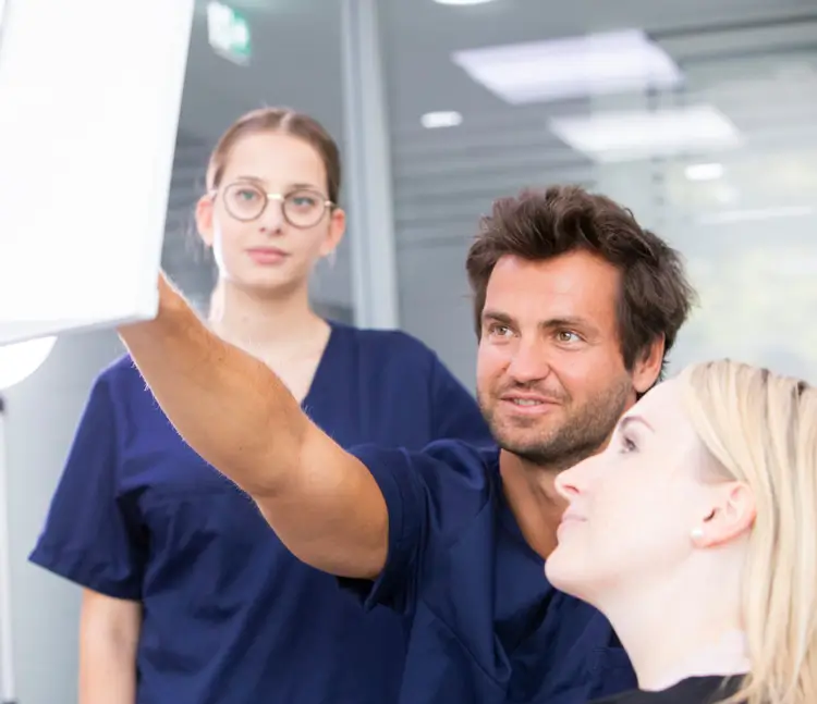 Zahnarztteam in blauen Kitteln betrachtet gemeinsam mit einer Patientin eine Aufnahme auf einem Screen