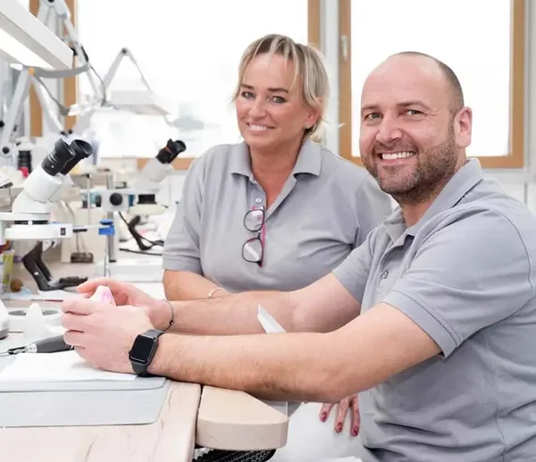 Zwei Labortechniker sitzen an ihren Arbeitspl&auml;tzen und l&auml;cheln in die Kamera