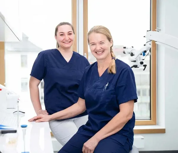 Zwei Zahnärztinnen in blauer Arbeitskleidung sitzen und lächeln in einer modernen Praxis mit großen Fenstern