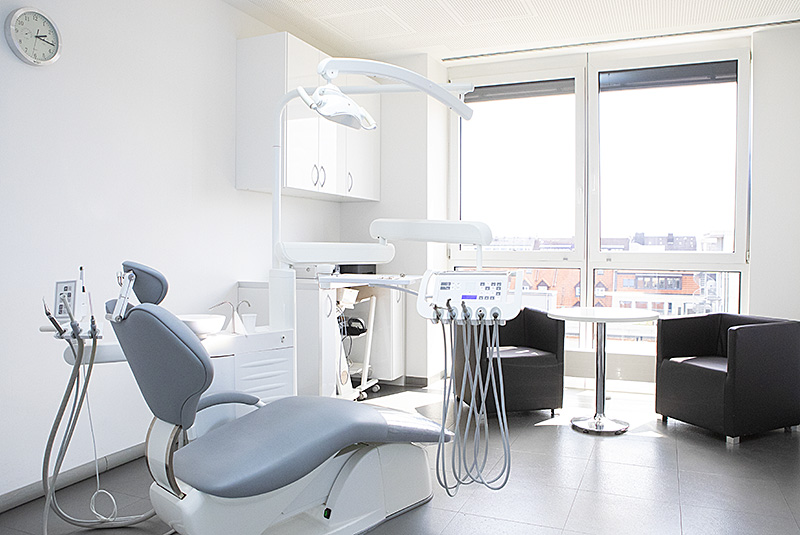 Behandlungszimmer mit Patientenstuhl, zwei grauen Sesseln und einem kleinen Tisch