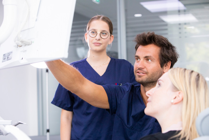 Eine Oralchirurgin und ein Oralchirurg zeigen einer Patientin einen Befund auf einem Röntgenbild